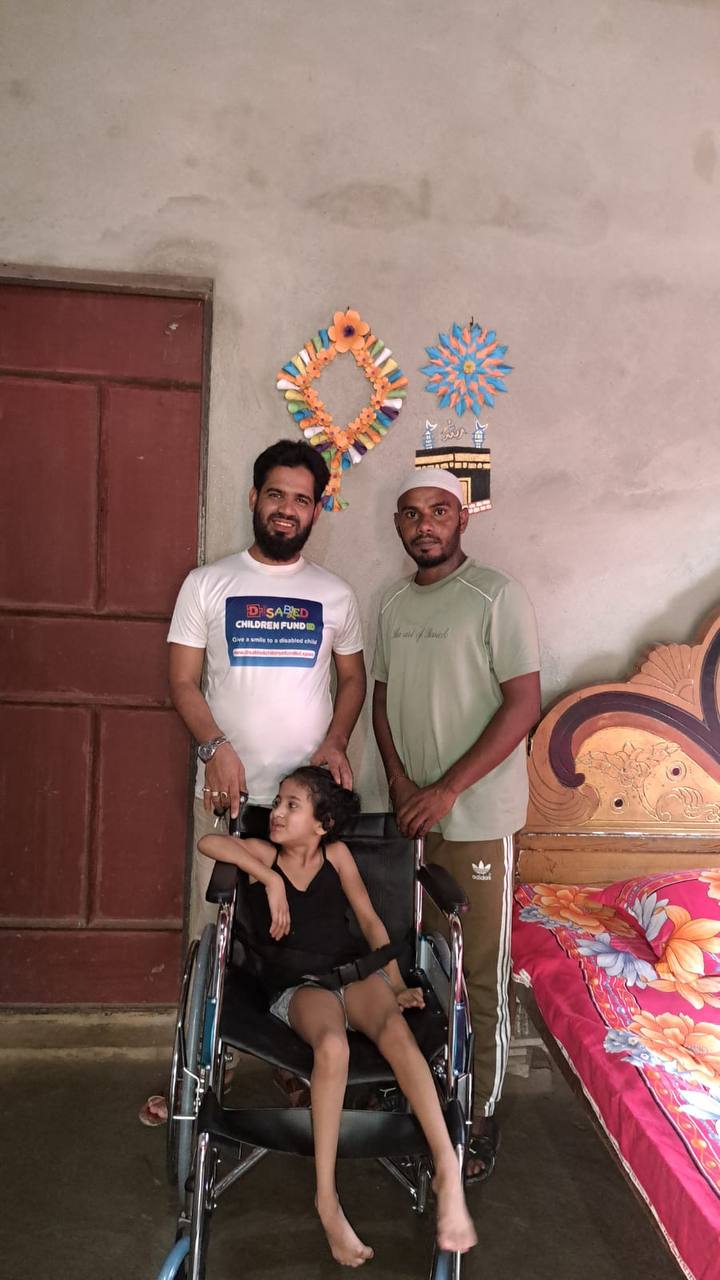 A disabled child in her new wheelchair, with her father and a Disabled Children Fund team member in their home in rural Bangladesh